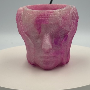 May include: A translucent pink and white marbled planter shaped like a human head with stylized wings on the sides. The surface has a glittery, textured finish with a hollow opening at the top, featuring a sculpted face with closed eyes.