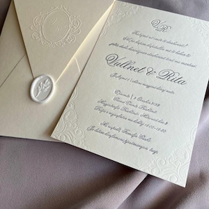 May include: Ivory wedding invitation suite with embossed floral details and a wax seal. The invitation features elegant script and the names "Vullnet & Rita". The envelope has a monogram and is sealed with a white wax seal.