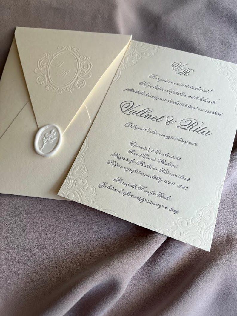 May include: Ivory wedding invitation suite with embossed floral details and a wax seal. The invitation features elegant script and the names "Vullnet & Rita". The envelope has a monogram and is sealed with a white wax seal.
