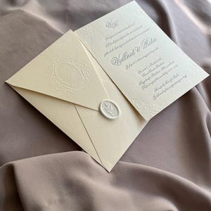 May include: Ivory wedding invitation suite with embossed floral details. The invitation features the names "Vullnet & Rita" and event details. The envelope is sealed with a wax stamp. The suite rests on a taupe fabric background.