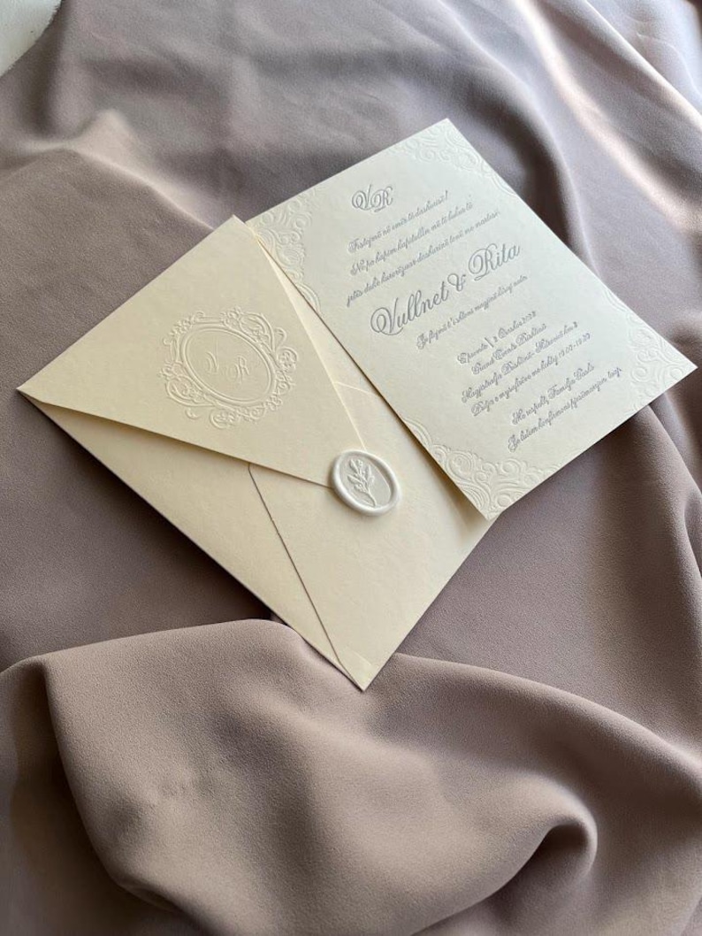 May include: Ivory wedding invitation suite with embossed floral details. The invitation features the names "Vullnet & Rita" and event details. The envelope is sealed with a wax stamp. The suite rests on a taupe fabric background.