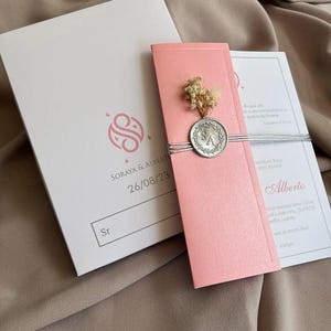 May include: A wedding invitation suite featuring a pink invitation card with a silver seal and dried flowers. The suite includes a white envelope with a pink logo and text, and a white card with text. The date 26/08/23 is visible.
