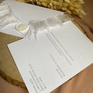 May include: White wedding invitation with a feather trim and a wax seal. The invitation includes the names "Ayşenur & Ali" and the date "08/06/2024".