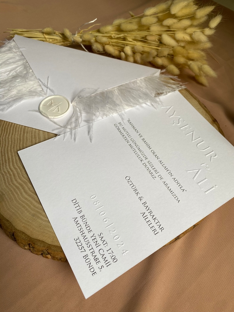 May include: White wedding invitation with a feather trim and a wax seal. The invitation includes the names "Ayşenur & Ali" and the date "08/06/2024".