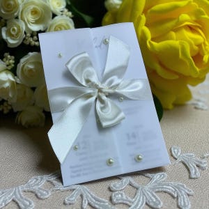 May include: A white wedding invitation card with a satin ribbon bow and scattered pearls. The card is open, revealing printed text and numbers. White roses and a yellow rose are in the background.