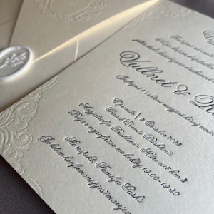 May include: Ivory-colored wedding invitation suite with embossed floral details and elegant script. Includes an envelope with a wax seal and a decorative border. The invitation text is in a foreign language.