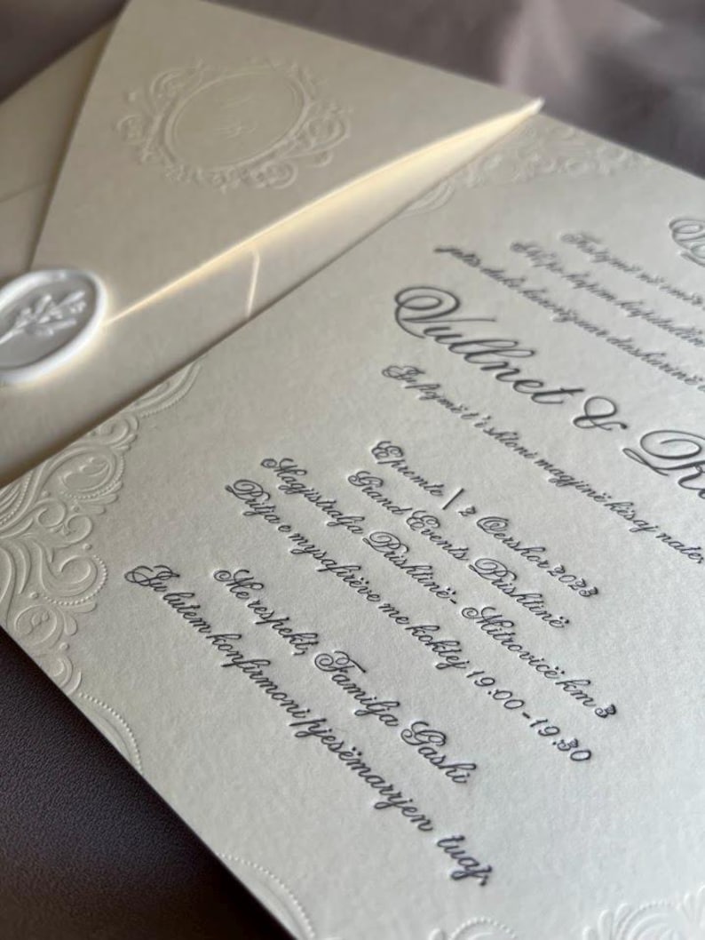 May include: Ivory-colored wedding invitation suite with embossed floral details and elegant script. Includes an envelope with a wax seal and a decorative border. The invitation text is in a foreign language.