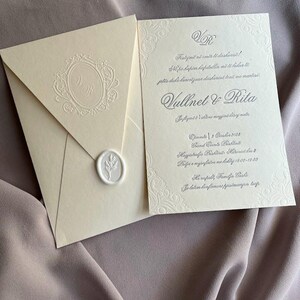 May include: Ivory wedding invitation suite with embossed details. The envelope features a monogram and a wax seal. The invitation card includes decorative borders and text in a script font. The suite is displayed on a mauve fabric background.