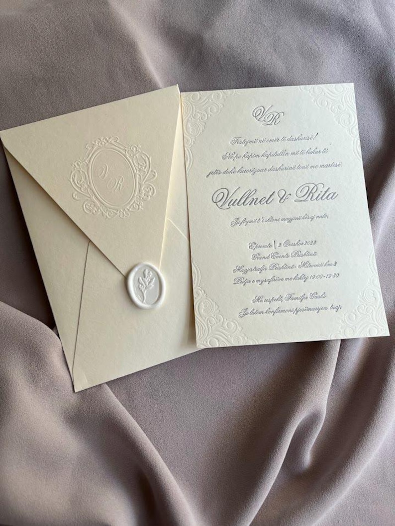 May include: Ivory wedding invitation suite with embossed details. The envelope features a monogram and a wax seal. The invitation card includes decorative borders and text in a script font. The suite is displayed on a mauve fabric background.