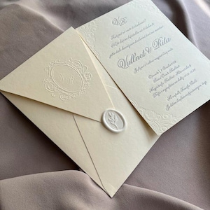 May include: Ivory wedding invitation suite featuring an envelope sealed with a wax stamp and a matching invitation card. The envelope has an embossed monogram and the invitation has decorative borders and text in a script font.