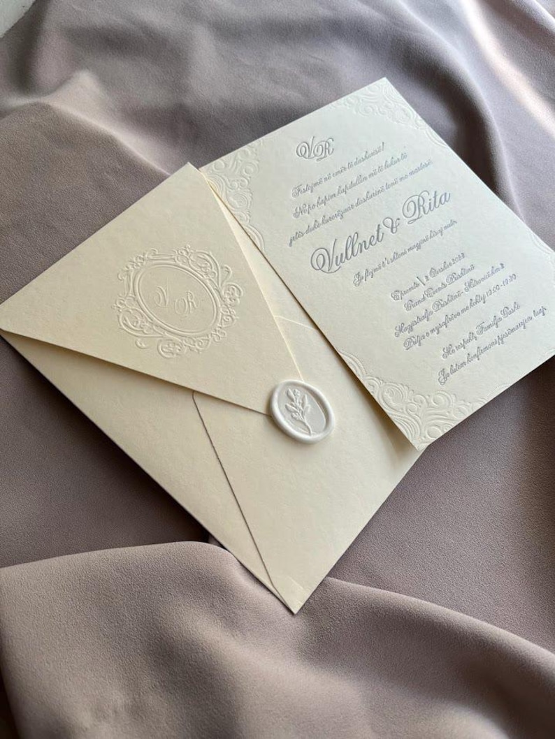 May include: Ivory wedding invitation suite featuring an envelope sealed with a wax stamp and a matching invitation card. The envelope has an embossed monogram and the invitation has decorative borders and text in a script font.