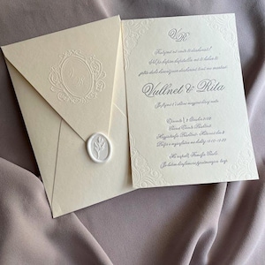 May include: Ivory wedding invitation suite with embossed details. The invitation features ornate borders and text, including names and event details. An envelope with a monogram and a wax seal is included.