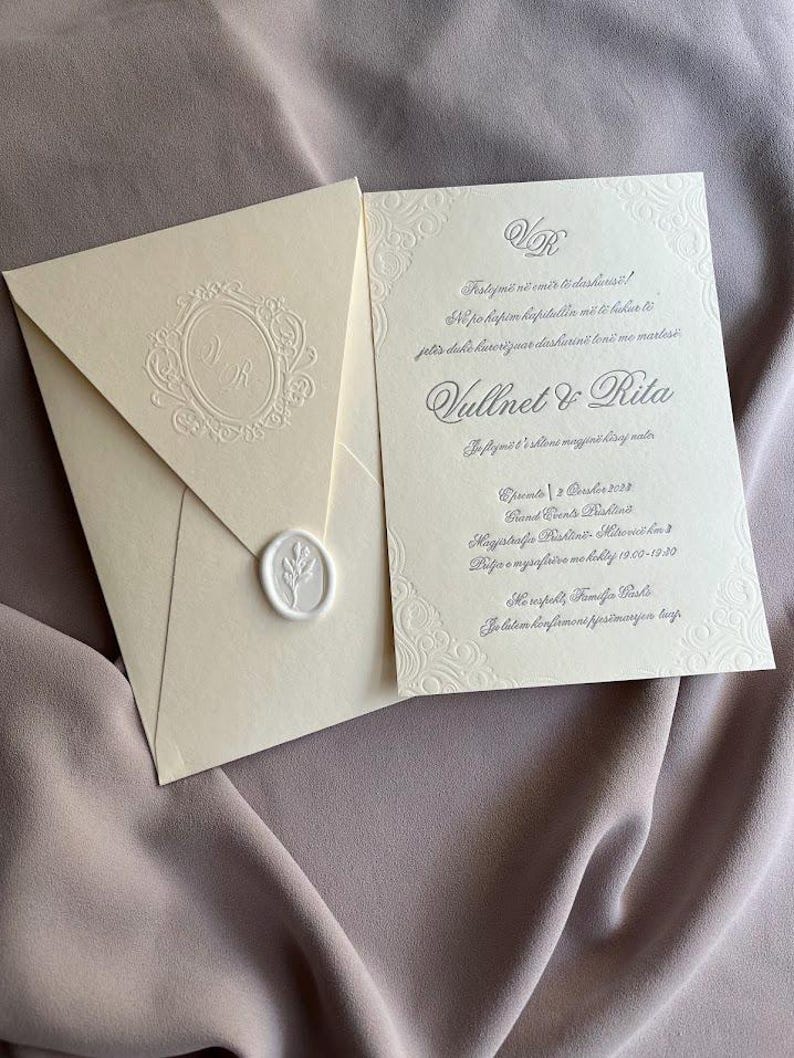 May include: Ivory wedding invitation suite with embossed details. The invitation features ornate borders and text, including names and event details. An envelope with a monogram and a wax seal is included.