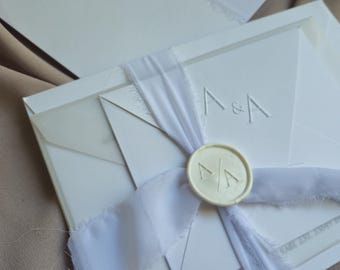May include: White wedding invitation with a white ribbon and a wax seal with the initials "A & A". The invitation is on a light brown surface.