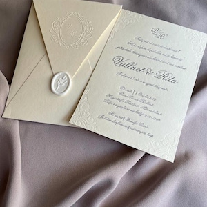 May include: Ivory wedding invitation suite with embossed details. The envelope features a monogram and a wax seal. The invitation card includes decorative borders and elegant script. The suite is displayed on a mauve fabric background.