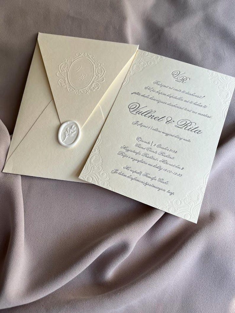 May include: Ivory wedding invitation suite with embossed details. The envelope features a monogram and a wax seal. The invitation card includes decorative borders and elegant script. The suite is displayed on a mauve fabric background.