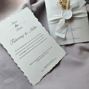 May include: Elegant wedding stationery set featuring a "Save the Date" card and an invitation wrapped with a white ribbon and a wax seal that says "Love". The cards have a textured, torn-edge design, with Greek text.