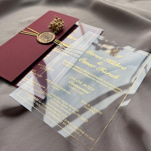 Invitación de boda de lujo en acrílico con detalles en lámina dorada, sobre color burdeos, sello de cera y diseño de acrílico transparente.