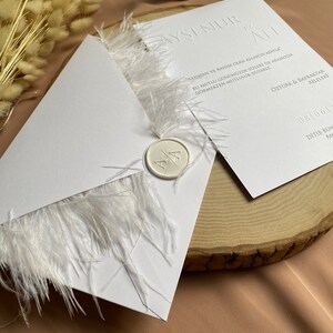 May include: White wedding invitation with a white feather trim and a wax seal with the initials "A" and "A". The invitation text reads "Ayşinur & Ali" and includes the date "08/06/" and the text "Ditib Bon" in Turkish.
