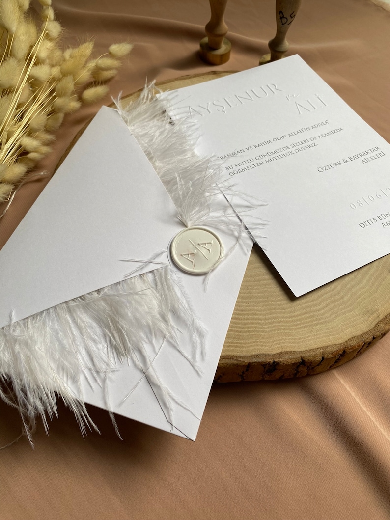 May include: White wedding invitation with a white feather trim and a wax seal with the initials "A" and "A". The invitation text reads "Ayşinur & Ali" and includes the date "08/06/" and the text "Ditib Bon" in Turkish.