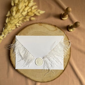 May include: A white envelope with a white feather trim and a white wax seal with the initials "M A" on a wooden slice. The envelope is on a light brown surface with a bouquet of dried pampas grass.