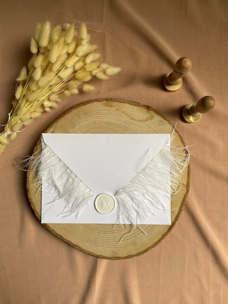 May include: A white envelope with a white feather trim and a white wax seal with the initials "M A" on a wooden slice. The envelope is on a light brown surface with a bouquet of dried pampas grass.