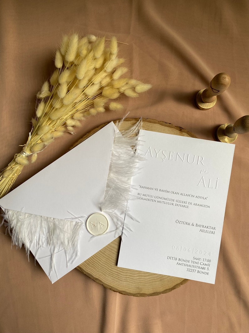 May include: White wedding invitation with a white feather trim and a wax seal. The invitation reads "Ayşenur & Ali" and includes the date "08/06/2024" and the time "17:00". The invitation also includes the location "Ditib Bunde Yeni Cami, Amthausstra&szlig;e 5, 32257 Bunde".
