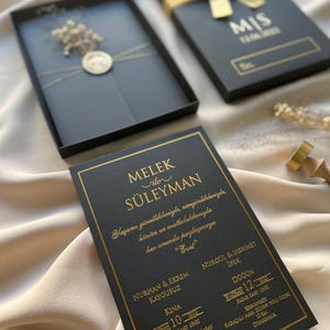 May include: Elegant black wedding invitation set with gold foil lettering. The invitation card features the names "MELEK ile SÜLEYMAN" and event details. The set includes a box tied with a gold ribbon and a matching envelope.