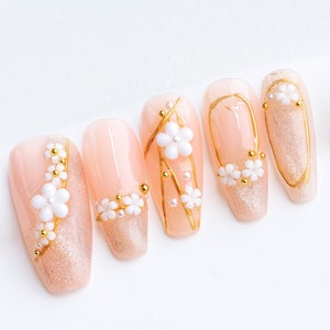 May include: Six nude-colored fake nails with gold accents and white 3D flowers. The nails are decorated with gold glitter and small white pearls.