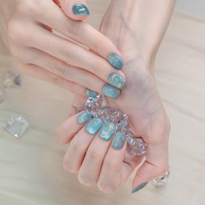 May include: Close-up of a manicure featuring short, oval-shaped nails. The nails are painted in a gradient of teal and blue, with gold leaf accents. The design includes small fish illustrations on some nails. The hands are holding a clear, faceted object.