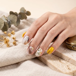May include: A hand with manicured nails in various designs. One nail is solid yellow, while others feature marble effects, gold accents, and floral patterns. The nails are oval-shaped and showcase a range of colors, including white, brown, and gold.