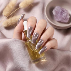 May include: A clear glass dropper bottle filled with a golden yellow liquid, held by fingers featuring long, coffin-shaped nails with a metallic, shimmering taupe cat-eye polish finish, showcasing a reflective light streak across each nail surface.