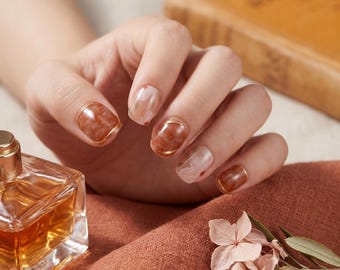 May include: Close-up of a hand with manicured nails. The nails feature a marbled brown and clear design with gold trim. A perfume bottle and a folded orange fabric are in the foreground. Dried flowers and a book are in the background.