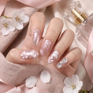 May include: Oval-shaped nails featuring a pearlescent pink base coat, decorated with white painted cherry blossom motifs, small white pearls, and silver butterfly-shaped charms. The nail art design incorporates delicate floral patterns.
