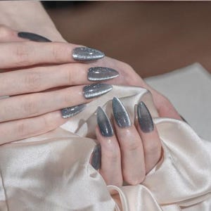 May include: Close-up of hands with long, pointed, gray and silver glitter nail polish. The nails have a cat-eye effect, with a gradient from dark gray to silver. The nails are on a satin fabric.