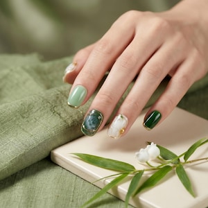 May include: Close-up of manicured nails in shades of green and white with gold accents. Some nails feature a solid colour, while others showcase intricate designs. The nails are set against a soft green backdrop, with a sprig of white flowers and green leaves.