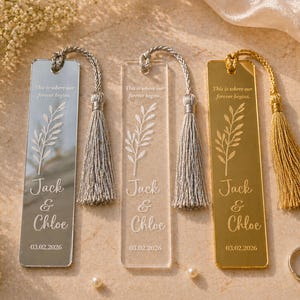 May include: Three personalized bookmarks with tassels. One is silver, one is clear, and one is gold. Each bookmark has a leaf design, the names "Jack & Chloe", and a date. Wedding rings and pearls are also in the image.