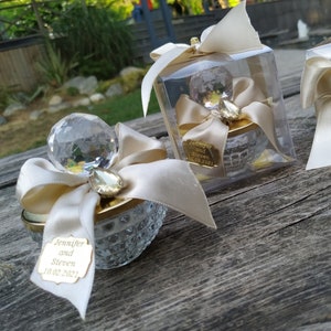 May include: Three small, clear glass containers with gold accents and a crystal-like top. Each container is decorated with a white satin ribbon and bow. One container has a tag with the text "Jennifer and Steven 10.02.2021".