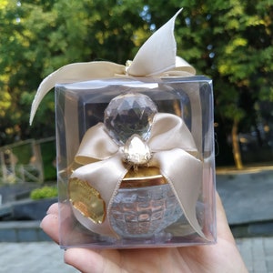 May include: A clear glass jar with a gold lid and a crystal-like top. The jar is decorated with a gold ribbon and a small crystal. The jar is in a clear plastic box with a white ribbon.