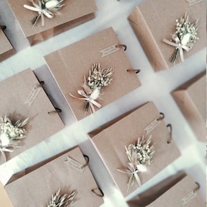 May include: Multiple brown paper gift bags with white and beige dried flower arrangements tied with a beige ribbon. The bags have a brown paper handle and a small tag with handwritten text.