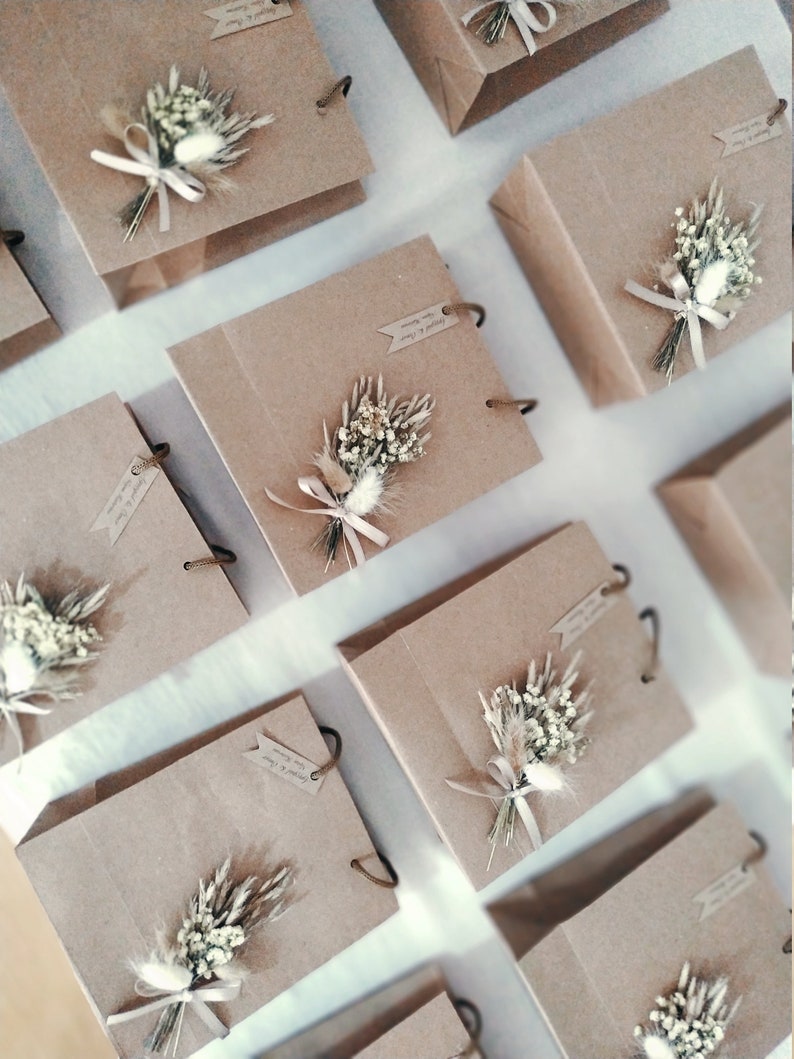 May include: Multiple brown paper gift bags with white and beige dried flower arrangements tied with a beige ribbon. The bags have a brown paper handle and a small tag with handwritten text.