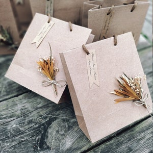 May include: A close-up of a brown paper gift bag with a gold and white dried flower arrangement. The bag has a tag