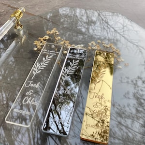 May include: Three personalized acrylic bookmarks with floral designs and names "Jack & Chloe" and a date. One is clear, one is mirrored, and one is gold. They rest on a clear tray with gold handles, with dried flowers.