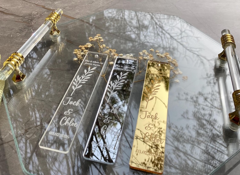 May include: Three personalized acrylic bookmarks with floral designs and names "Jack & Chloe" and a date. One is clear, one is mirrored, and one is gold. They rest on a clear tray with gold handles, with dried flowers.