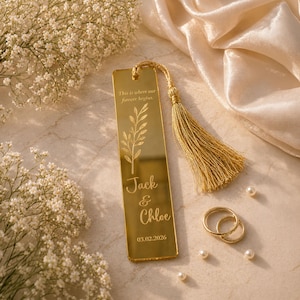 May include: A gold-colored bookmark with a tassel, engraved with the names "Jack & Chloe" and the date 03.02.2026. The bookmark features a floral design and the text "This is where our forever begins." Two gold rings and pearls are also present.