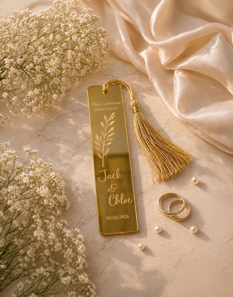 May include: A gold-colored bookmark with a tassel, engraved with the names "Jack & Chloe" and the date 03.02.2026. The bookmark features a floral design and the text "This is where our forever begins." Two gold rings and pearls are also present.