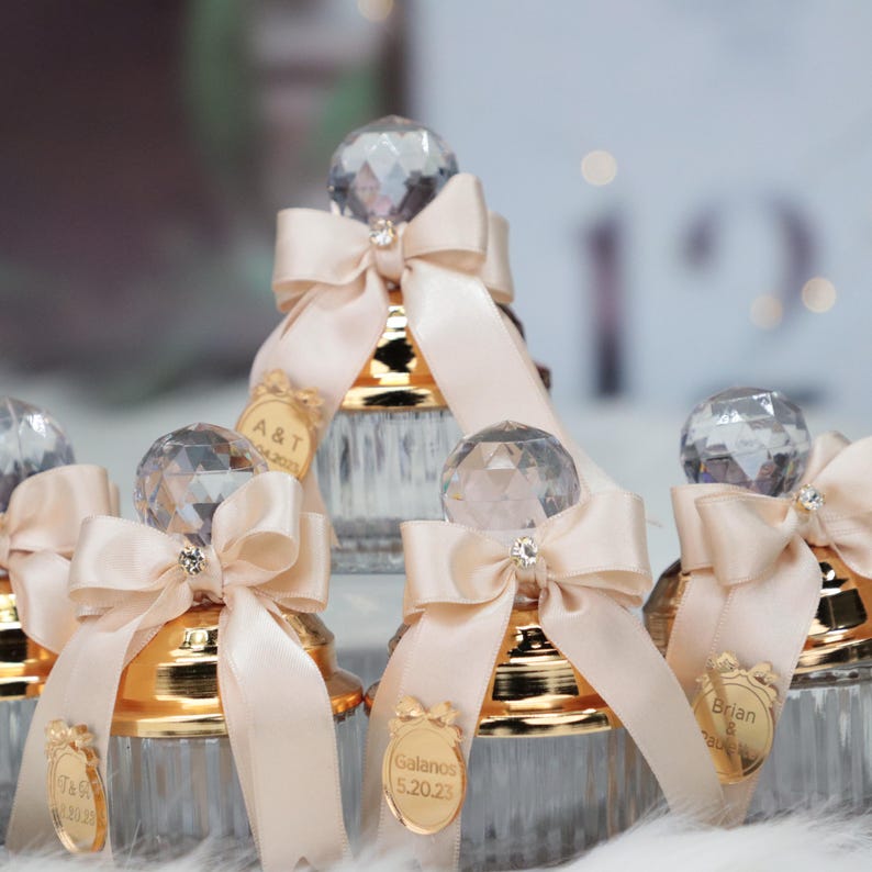 May include: Set of four clear glass jars with gold lids and crystal-like tops. Each jar has a beige satin ribbon tied around the top and a personalized gold sticker with a name and date.