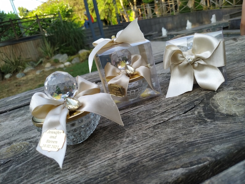 May include: Three small glass containers with crystal tops and gold accents. Each container is decorated with a white satin bow and a small crystal. One container has a personalized tag with the text "Jennifer and Steven 10.02.2021".