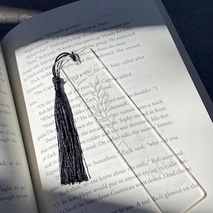May include: Clear acrylic bookmark with a black tassel, resting on an open book. The bookmark features an etched leaf design and the words "Zack & Inky 2026". A stylish accessory for book lovers.