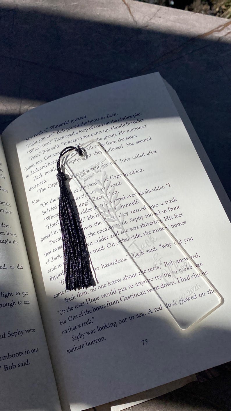 May include: Clear acrylic bookmark with a black tassel, resting on an open book. The bookmark features an etched leaf design and the words "Zack & Inky 2026". A stylish accessory for book lovers.